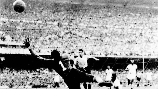 A imagem em preto e branco mostra um lance de gol em uma partida de futebol, com a bola já ultrapassando o alcance do goleiro, que salta em vão em direção ao canto superior da meta. Ao fundo, jogadores acompanham a jogada diante de um estádio lotado, em um registro histórico que destaca o momento decisivo da final.