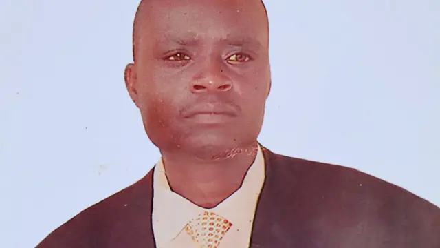 Photographie d'un instantané de Samuel Owino Owoyo. Il a l'air sérieux dans sa veste sombre, chemise blanche et cravate à pois.