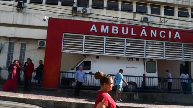 Fachada de hospital com letreiro 'Ambulância' e pessoas em volta