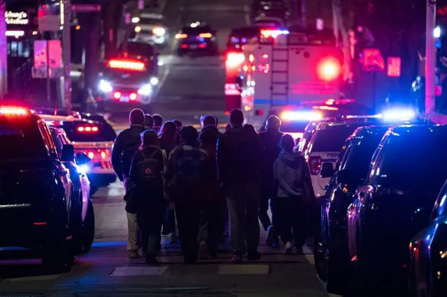 Polis, bir üniversite binasından tahliye edilen öğrencilere eşlik ediyor. Öğrenciler sokakta toplanmış vaziyette. Etraflarında polis arabaları var.