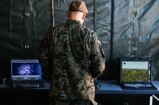 Польський військовий стоїть спиною до камери. Він дивиться на два екрани моніторів, де видно проліт дронів 