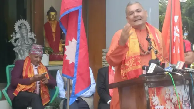दुर्गा प्रसाई बोल्दै गर्दा पृष्ठभूमिमा मोबाइल फोन हेर्दै बसेका धवलशमशेर राणा