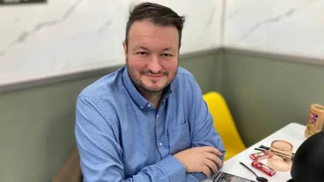 David, sentado a una mesa, con camisa azul, barba oscura y cabello corto y oscuro, sonríe y mira a la cámara.