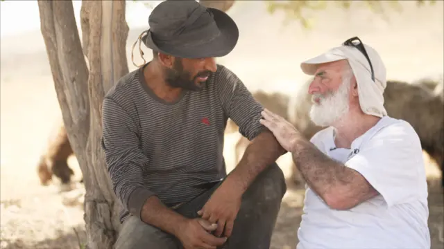 احمد، کشاورز فلسطینی که یک بلوز آستین بلند پوشیده و کلاهی بر سر دارد، در کنار گیل نشسته که ریش سفیدی دارد. گیل دست خود را روی بازوی احمد گذاشته و این دو که زیر یک درخت نشسته‌اند، با هم در حال حرف زدن است.