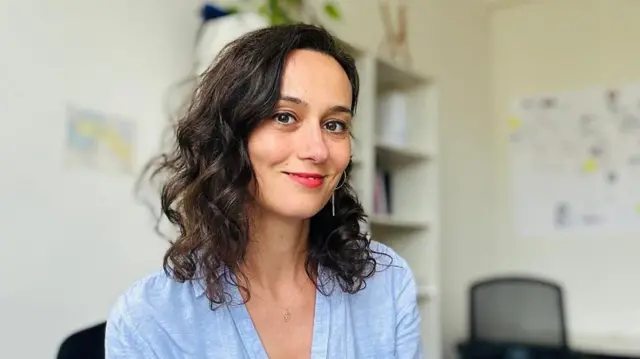 İrem mavi gömlekli ofiste kameraya bakıyor. 