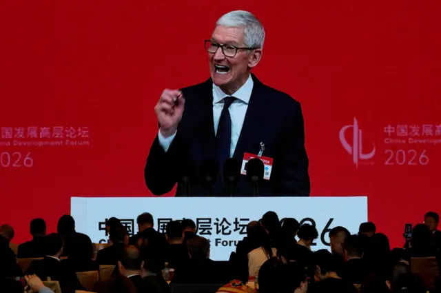 Кук на большом экране, перед которым спиной к фотографу сидят люди. Задний план красного цвета