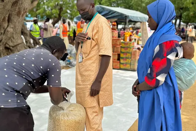 Aisha Abubakar (à droite), vêtue d'une robe bleue sur une chemise à motifs bleu marine, rouge et blanc, fait la queue avec son bébé de sept mois sur le dos derrière un homme portant un masque facial et une chemise longue beige et un pantalon pour obtenir un sac de maïs, qui est en train d'être ficelé par un fonctionnaire à Gwoza, au Nigeria, en juillet 2025.