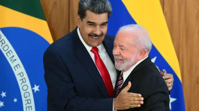 A imagem mostra dois homens se abraçando e sorrindo durante um encontro oficial, tendo ao fundo bandeiras do Brasil e da Venezuela. Um deles segura uma caneta na mão enquanto ambos posam em um gesto amistoso diante das câmeras.