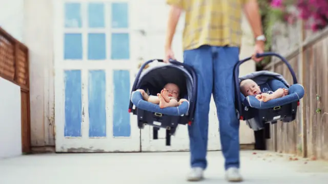 Imagem de uma pessoa segurando duas cadeirinhas de carro para bebês com bebês sentados nelas. 