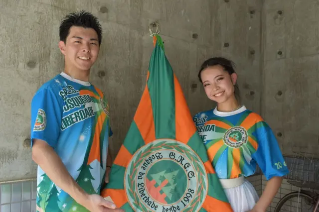 A foto mostra dois jovensroleta de verdadeaparência oriental, pele clara, com o uniforme da escolaroleta de verdadesamba que é azul, laranha e verde, e segurando uma bandeira entre eles.