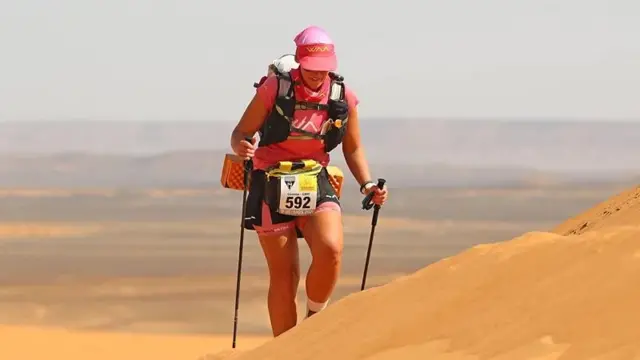 Uma mulher caminhando em um deserto com apoio. Ela usa um número de identificação em uma competição.