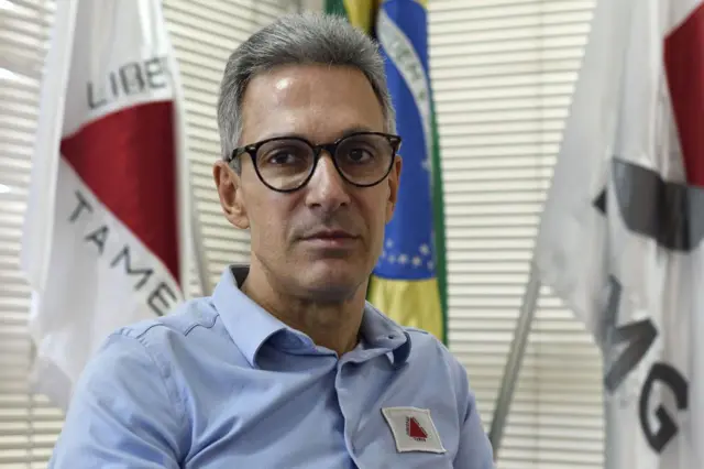 Romeu Zema é um homem branco e usa óculos de grau com armação preta. Ele está com uma camisa azul clara que tem uma bandeira de Minas Gerais. 