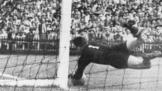 A imagem em preto e branco mostra um goleiro mergulhando junto à trave para defender uma bola rente ao chão durante uma partida de futebol. Ao fundo, uma multidão acompanha o lance das arquibancadas, em um registro histórico que destaca a intensidade e o dramatismo do momento.