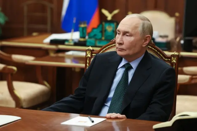 Vladimir Putin koltukta oturuyor, arkasında Rus bayrağı görünüyor