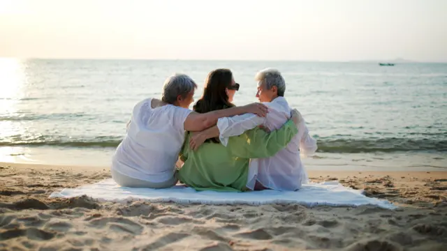 तीन महिलाएं बीच पर एक दूसरे के कंधे पर हाथ रखे बैठी हैं