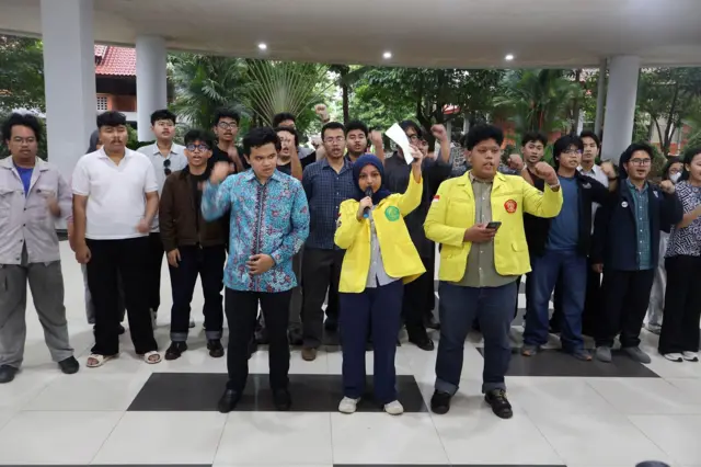 Sekelompok mahasiswa UI sedang mengngkat tangan kiri mereka di sebuah pelataran. Paling depan terdapat seorang mahasiswa berhijab dan beralmamater kurning di tengah, diapit seorang pria dengan almamater yang sama dan pria berbaju batik.
