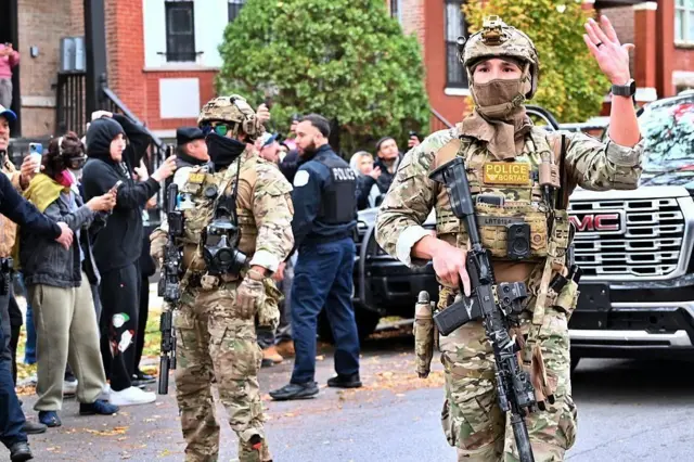 道路に大きな黒い車があり、その近くに迷彩服を着て自動小銃をもった当局者や、背中にPOLICEの文字が入ったベストを着た人が立っている。道路の端には人々が横に並び、スマホで取り締まりの様子を撮影するなどしている