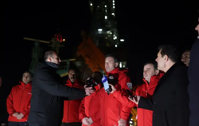 Bayraktar ve ekibi kırmızı montlarla, Bakan basına açıklama yapıyor, iki muhabir mikrofonları tutuyor, gece. 