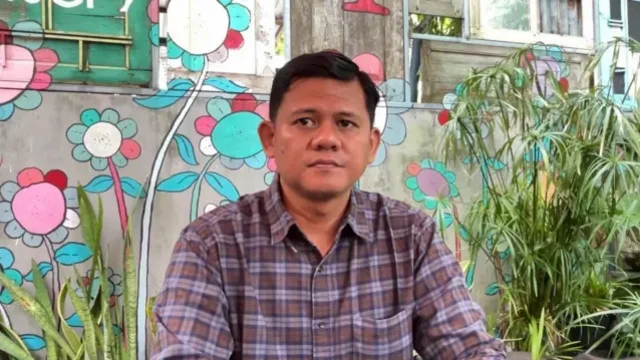 A man in a flannel button-up shirt sitting in front of a wall painted with colourful flowers.