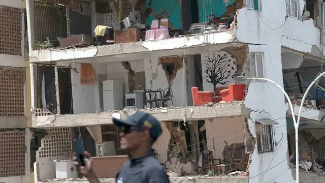 Un hombre está parado frente a un edificio que perdió su fachada, dejando al descubierto muebles y habitaciones en habitaciones con paredes dañadas o destruidas
