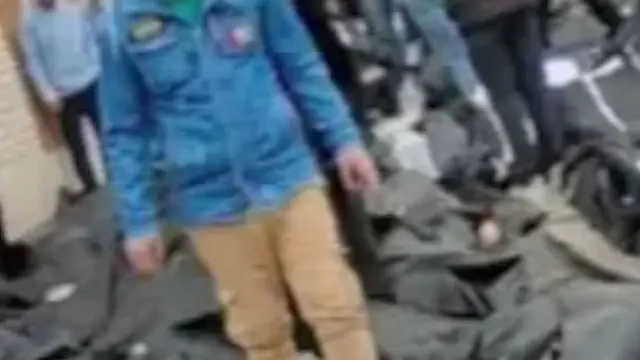 Des corps gisent sur le sol dans des sacs noirs tandis qu'un homme marche à proximité. D'autres personnes se tiennent à proximité. Personne n'est identifiable sur l'image.