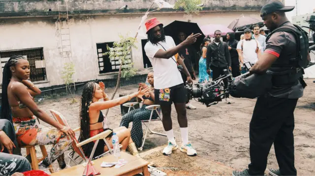 Meji Alabi, o de fila pupa, o wọ aṣọ funfun, ṣokoto kekere dudu ati ibọsẹ ati bata funfun. O gbe kamẹra dani, na ọwọ rẹ siwaju bo ṣe n dari iṣẹ, awọn obinrin kan jokoo, bẹẹ ni awon eeyan kan naa duro ninu aworan yii.