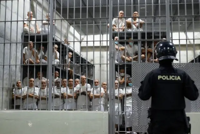 Presos com uniformes brancos estão na cela de uma prisão enquanto são observados por um policial