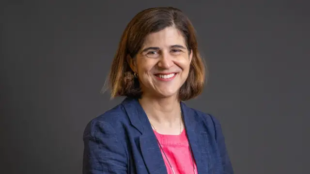 Sarah Sackman aparece en la foto sonriendo a la cámara frente a un fondo oscuro y liso, vistiendo un blazer sobre una blusa roja.