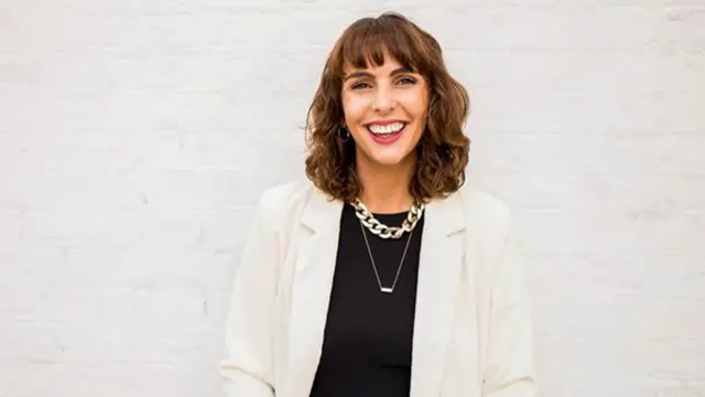 Знімок Вікі Павітт у білому костюмі з чорною блузкою, вона має хвилясте каштанове волосся до плечей і усміхається в камеру