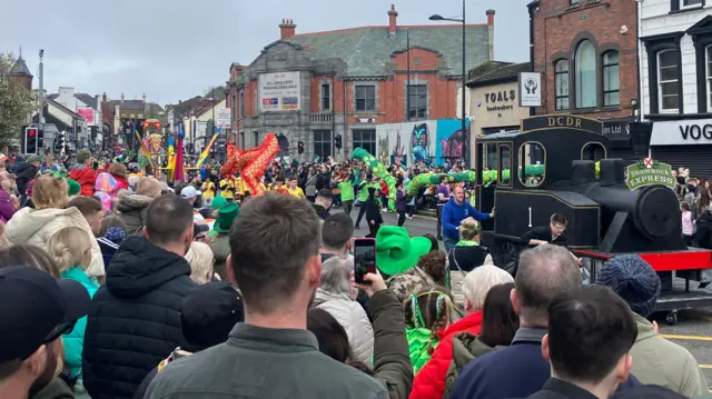 A bustling crowd, don in festive green attire and hats, lines a city street to watch a vibrant St. Patrick Day parade. Central to di scene is a float resembling a black steam locomotive wey dey labeled 'St. Patrick's Express,' to promote di Downpatrick & County Down Railway. Di parade route dey flanked by various buildings, including shops like 'VOGUE' and 'O2.' 