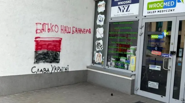 На стіні недбале графіті: вгорі червоним кольором написано «Батько наш Бандера», під ним намальований червоно-чорний прямокутний прапор, а нижче чорним кольором написано «Слава Україні». Праворуч видно вхід до медичного магазину з вітриною та скляними дверима, на яких розміщені наліпки та інформаційні таблички польською мовою