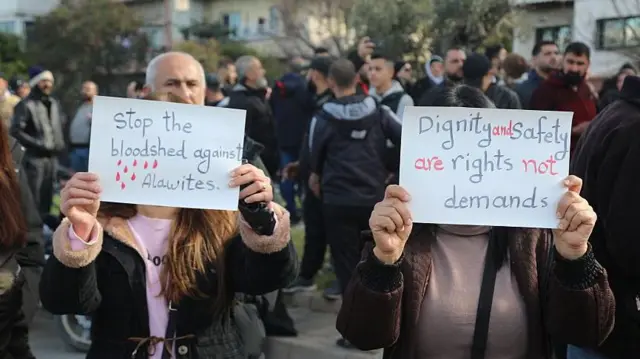 Толпа из примерно 20 протестующих, на переднем плане две женщины, закрывающие лицо плакатами. На них написано «остановите резню алавитов» и «достоинство и безопасность — это права, а не требования».