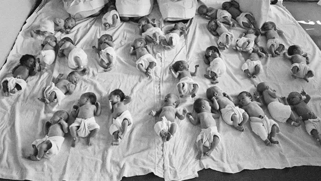 Um grupo de bebês colocados juntos no chão de um hospital do NHS na década de 1960. Os bebês estão todos em um grande lençol branco e virados em direções diferentes, deitados no chão.