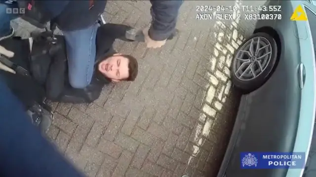 На парковці біля автівки, на спині лежить чоловік, намагаючись чинити опір поліції. Згори його руку тримає правоохоронець, одночасно притискаючи до землі коліном.