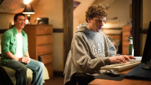 Jesse Eisenberg, con un buso gris de Gap, sentado frente a un computador con una cerveza al lado y un a migo mirándolo detrás.