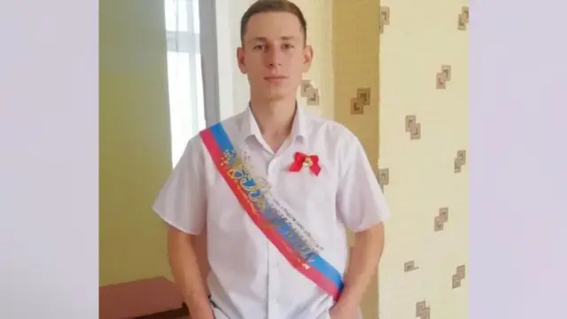 A picture of a young Russian man in a short-sleeved white shirt with a Russian strap over his shoulder