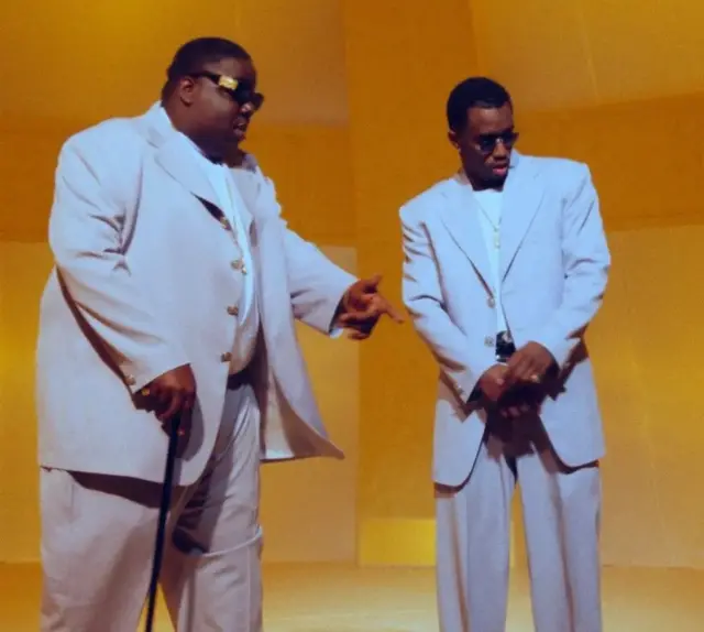 The Notorious B.I.G. (à esquerda) e Puff Daddy no set do videoclipe de "Hypnotize" em 1997. Ambos vestem branco e óculos escuros e estão em um cenário amarelo