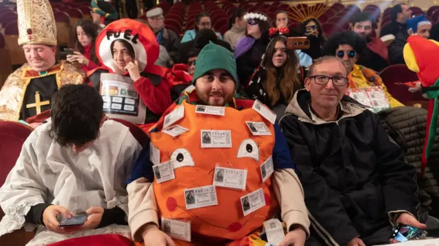 Los asistentes al sorteo en vivo disfrazados de obispo, estación de gasolina y de naranja.