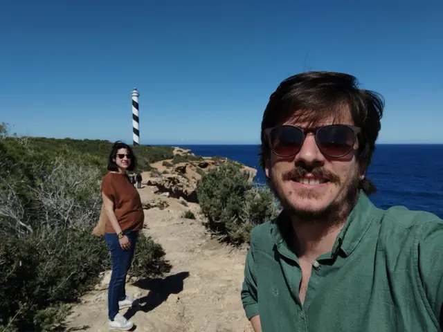 Almudena, de fondo, embarazada y Pablo, en primer plano. Es un selfie donde se les ve caminando por un sendero de piedras al lado del mar.