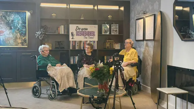 Ann a la izquierda, Middle Maureen y Doreen a la derecha. Tres ancianas estaban sentadas en sillas de ruedas, cubiertas por mantas grises. Tenían micrófonos manos libres. Detrás de ellas había una estantería con un cartel que decía "¿Recuerdas aquella vez?". Vemos una cámara montada en un trípode que las graba mientras hablan.