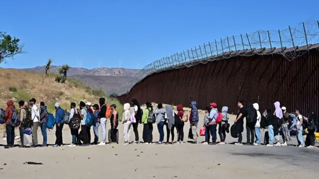 Migrantes hacen fila con la esperanza de ser procesados ​​por agentes de Aduanas y Protección Fronteriza después de que grupos llegaran a Jacumba Hot Springs, California, tras caminar bajo un calor intenso desde México hacia Estados Unidos