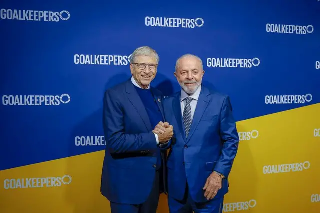 Gates e Lula sorrindo e apertando as mãos em frente a painel de evento