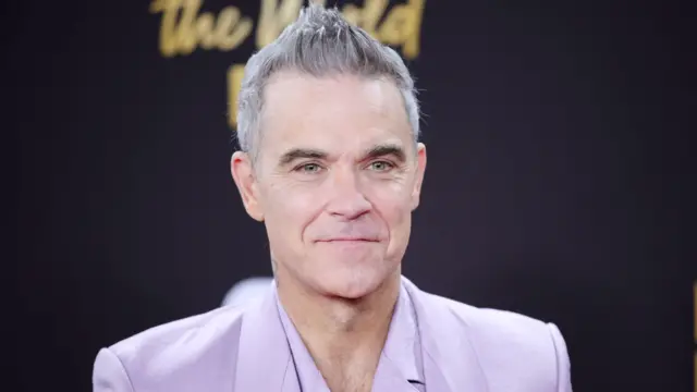 Robbie Williams, vestido con un traje color lila, sonríe mientras permanece frente a un fondo negro.