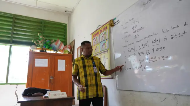 Rasyid mengajar di depan kelas sambil menerangkan huruf-huruf Hangeul yang dia tulis di papan tulis.