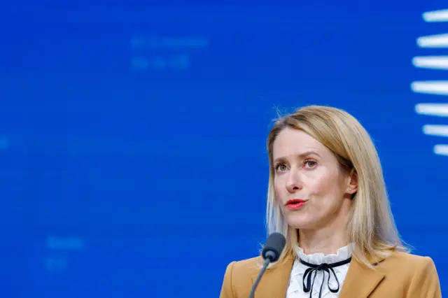 欧盟外交政策主管卡拉斯身穿米色外套,内搭白色黑蕾丝上衣,对着麦克风发言,背景为亮蓝色并带有白色图案元素