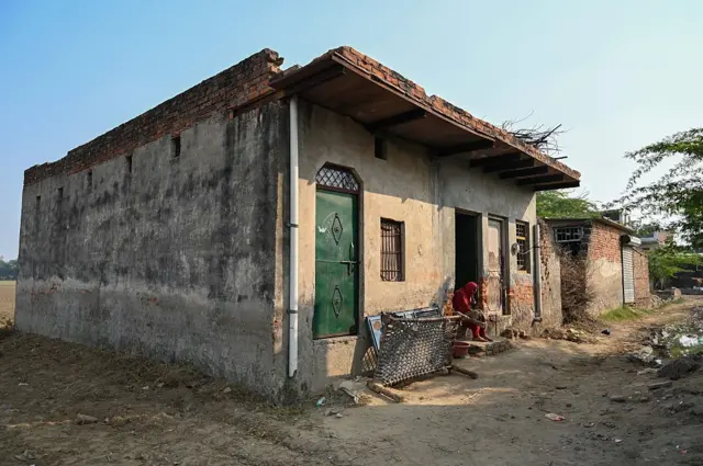 একটি এক তলা ঘর, তাতে সবুজ রংয়ের একটি দরজা লাগানো। অন্য একটি দরজায় লাল জামা পরা একজন নারী বসে আছেন। পাশে একটি বেতের বিছানা আড়াআড়ি উঠিয়ে রাখা। 