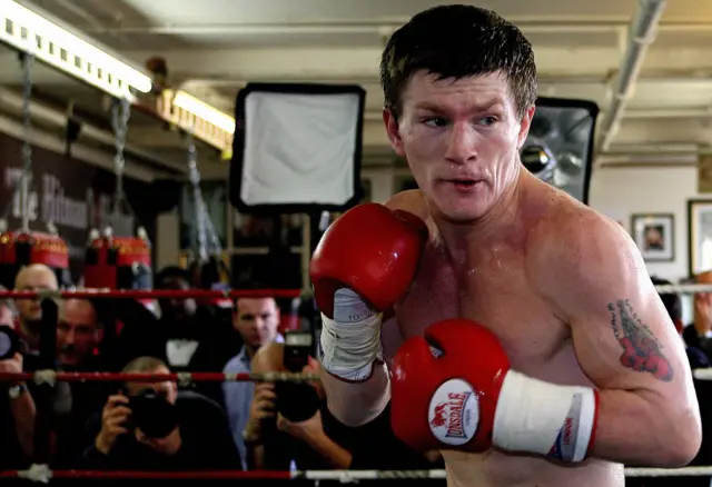 Ricky Hatton is inside a boxing ring wearing bright red gloves and white hand wraps. The boxer is shirtless, showing a tattoo on the upper arm. The stance is forward-facing with fists raised in a defensive position. In the background, there are punching bags, photographers, and lighting equipment, indicating a training session or media event in a gym setting.