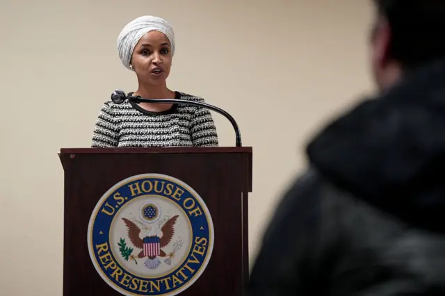 Ilhan Omar, kürsüde konuşma yapıyor, sırtı dönük bir erkek ekran önünde görünüyor