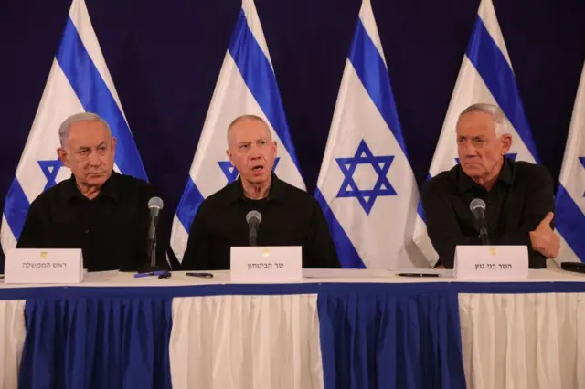 Benjamin Netanyahu, Yoav Gallant e Benny Gantz sentados numa mesa cercados por bandeirasverde casinoIsrael