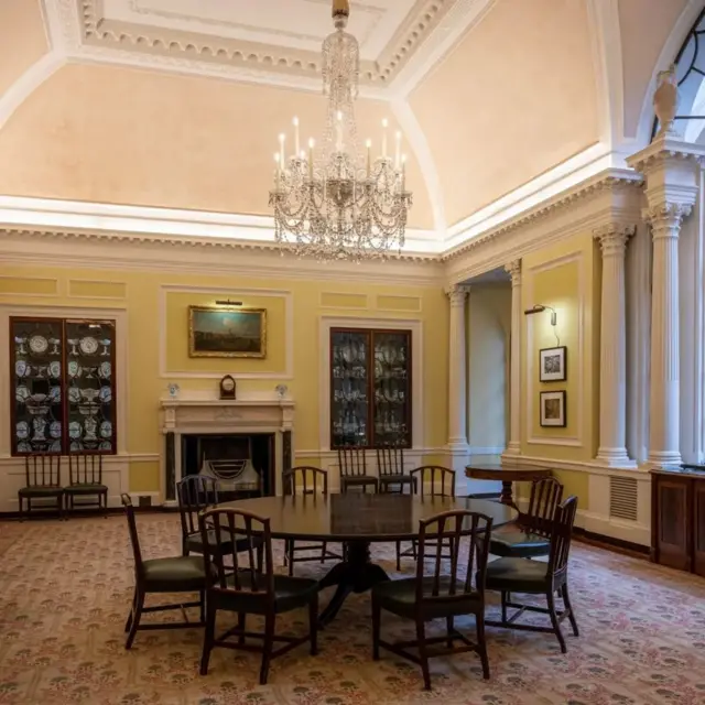 یک میز گرد با صندلی‌ها، دیوارهای زرد رنگ، اشیای زینتی قدیمی روی دیوار دیده می‌شوند